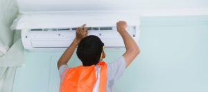 Technician installing an air conditioning unit on a wall, wearing an orange safety vest, in a light-colored room, highlighting HVAC installation services.