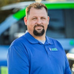 Man in blue polo shirt smiling, standing in front of a vehicle with green and blue branding, representing a professional service.