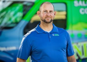Man in blue polo shirt standing in front of a brightly colored service vehicle, showcasing company branding and a professional demeanor.