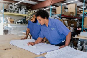 Two HVAC technicians reviewing blueprints on a table in a warehouse, showcasing teamwork and planning for Diamond Air Design services.